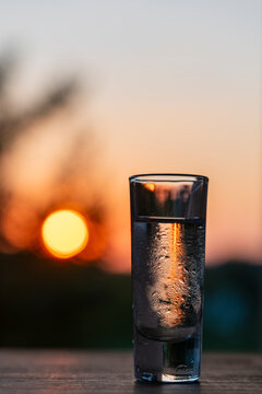 Cold vodka shot glass with condensation at sunset background. Closeup misty frosted shot glass filled with transparent alcohol liquor or vodka on wooden table against beautiful warm blurry sunset sky