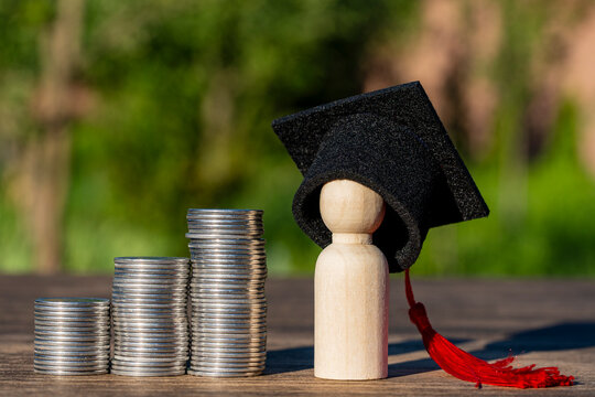 Wooden peg doll in graduation cap next coin stacks. Education savings concept with small wooden figure wearing black academic graduation hat with red tassel standing by stacks of silver coins
