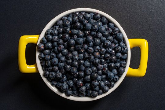 Fresh wild blueberries in yellow handles ceramic pot. Top view of ripe organic sweet blueberries filling a white ceramic saucepan with bright yellow handles placed on black textured background