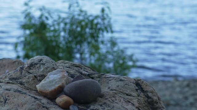 Smooth stones rest on large boulder beside blurred mountain lake water, creating a natural shoreline closeup with textured rock, soft blue waves and green vegetation in the background