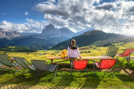 Woman hiker sitting on wooden bench looking at mountain view in Dolomites, Italy. Summer alpine resort landscape with colorful deck chairs, green grass meadow, yellow backpack and cloudy sky at sunset