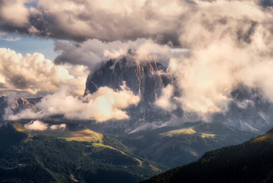 Rocky mountain peak surrounded by thick clouds and fog at sunset in Dolomites, Italy. Majestic alpine sunset landscape with dramatic cloudscape, sunbeams, high rocky cliffs and forest valley at dusk