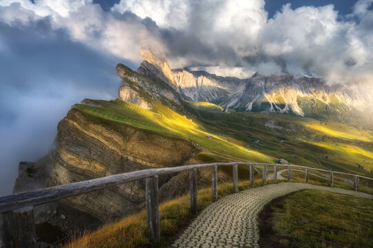 Seceda mountain peak in clouds with hiking trail and wooden fence in Dolomites, Italy. Summer alpine landscape with sunbeams, green grass meadow slope, high cliffs, dramatic sky and fog at sunset