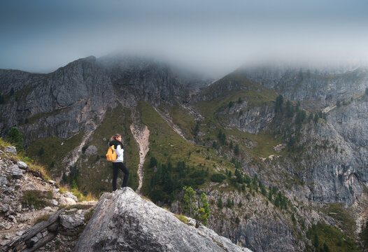 Woman hiker with yellow backpack standing on rock looking at mountain peaks in Dolomites, Italy. High altitude alpine landscape with girl, fog, clouds, rocky cliffs, forest slope and overcast sky