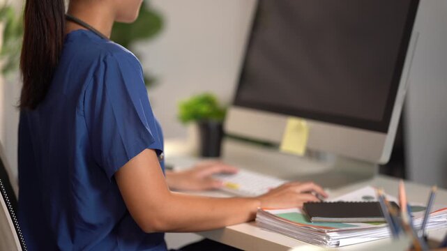 Focused Professional at Work: A dedicated woman in a blue uniform is engrossed in her work, typing on a computer in a bright office setting, surrounded by documents and office supplies.