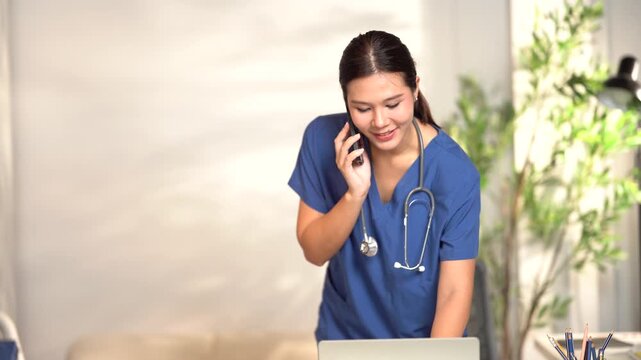 Nurse on Call and Computer: A female nurse in blue scrubs wearing a stethoscope is talking on the phone while working on a laptop in a bright, modern office environment with a plant in the background.