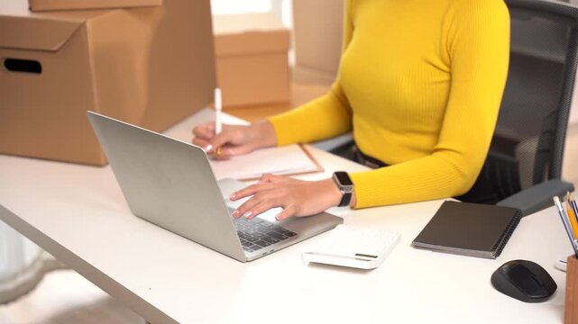 Online Seller at Work: A woman in a yellow shirt is working on her laptop in front of stacks of boxes. She is writing on a notebook while looking at her laptop, possibly managing her online store.