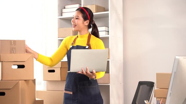 Small Business Owner Arranging Packages: A young woman, an entrepreneur, diligently organizes cardboard boxes, laptop in hand, in her workspace, ready for shipment.