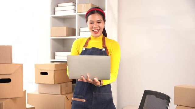 Joyful Entrepreneur with Laptop: A smiling young woman, wearing an apron, looks excitedly at her laptop surrounded by stacked cardboard boxes, embodying the spirit of a small business owner.