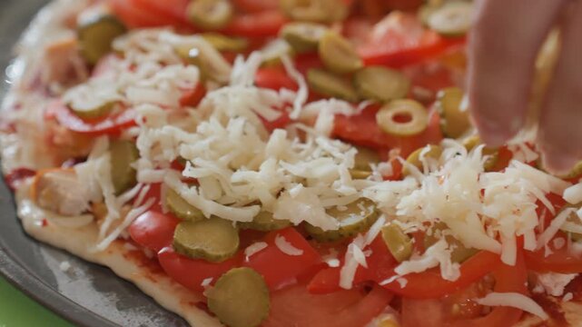 Overhead close view of rectangular pizza on metal tray, covered with tomato slices and green olive rings while shredded cheese is scattered across surface, showing savory meal preparation