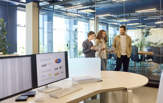 Colleagues in office meeting discuss business strategy. Three coworkers stand by glass boardroom reviewing reports, analytics on desktop screens. Clear concept of teamwork and strategy.