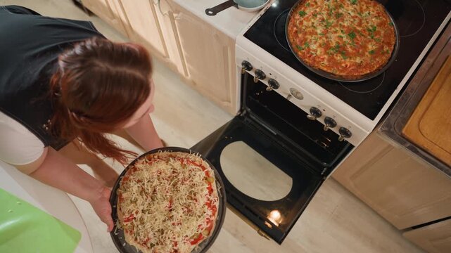 Overhead kitchen scene showing woman leaning forward to remove hot pizza pan from open oven and set it on counter while second pizza stays on stovetop, with warm indoor light and light wood floor
