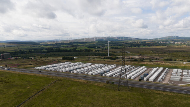 Aerial view of BESS battery energy storage system units with wind turbines in the background across a rural landscape in Lanark, Scotland, United Kingdom.