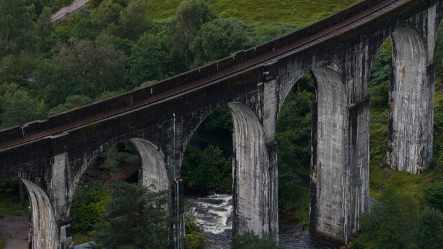 Aerial view of the historic Glenfinnan Viaduct spanning the lush landscape with a river flowing beneath its concrete arches in Glenfinnan, Scotland, United Kingdom.