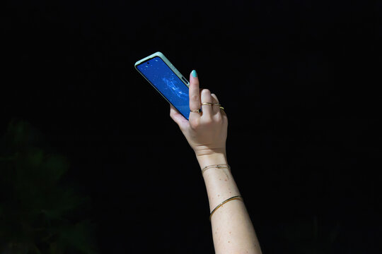 A raised arm holding a smartphone displaying a constellation map app against a completely black night sky, with jewelry visible on the wrist and fingers.