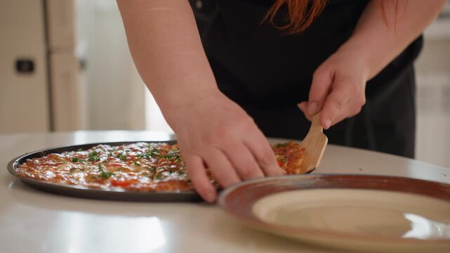 Close-up view of cook hands transferring freshly baked pizza slice from tray onto plate in warm kitchen light, with cheese stretch, slight bend, visible toppings and careful serving motion