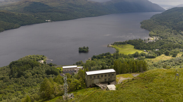 Aerial view of the Loch Sloy Hydro-Electric Scheme power station buildings nestled among lush green hills beside the dark waters of Loch Sloy in Arrochar, Scotland, United Kingdom.