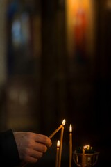 Naklejka premium Hand lighting a candle beside burning candles in a dark interior. Spiritual ritual, remembrance, prayer, and solemn memorial concept with atmospheric copy space.