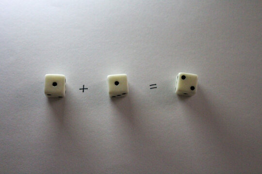 An addition math problem shows white dice &mdash; displaying numbers via their dots &mdash; on a white background, casting shadows. one plus one equals two