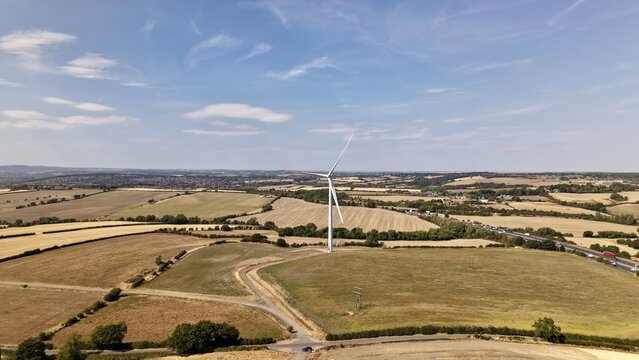 Aerial view of Penny Hill Wind Farm featuring a single wind turbine standing in a rural landscape with harvested fields, hedgerows, and a nearby road in Aston, England, United Kingdom.