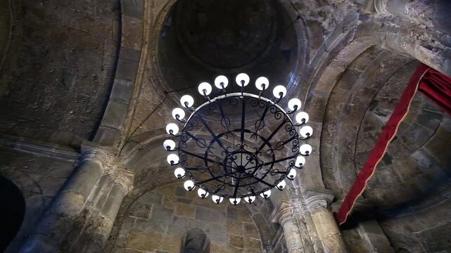 Interior low angle view of ancient stone Armenian apostolic church showing round metal chandelier, vaulted stone arches, columns, and dome under dark ceiling.