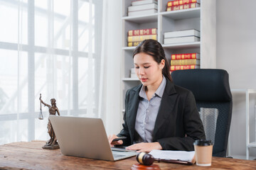Fototapeta premium Lawyer Working in Office: A focused female lawyer in a professional suit works diligently on her laptop at a wooden desk, surrounded by legal books and a statue of justice.