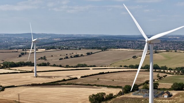 Aerial view of Penny Hill Wind Farm with large white wind turbines standing in golden harvested fields under a blue sky in Aston, England, United Kingdom.
