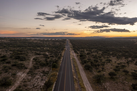 Aerial view of a straight road cutting through the rural landscape at sunset with scattered trees and a dramatic sky in Aguascalientes, Aguascalientes, Mexico.