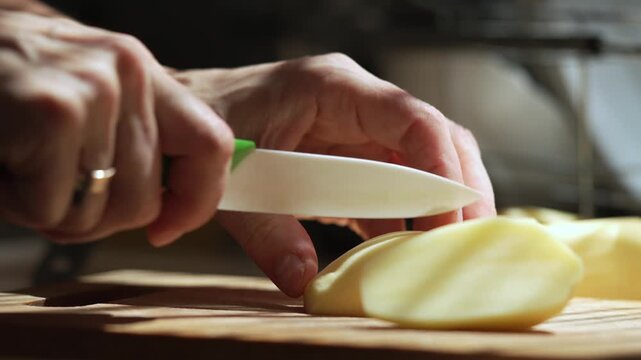 Hands Cutting Fresh Potato Slow Motion. Close up of hands cutting peeled fresh potato with a kitchen knife on a wooden cutting board in slow motion.