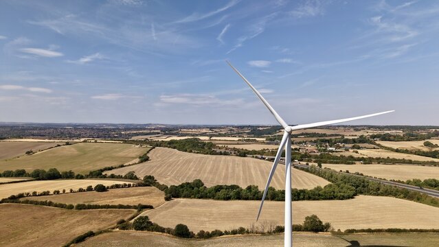 Aerial view of Penny Hill Wind Farm with a large wind turbine set against a rural landscape of golden fields and a clear blue sky in Aston, England, United Kingdom.