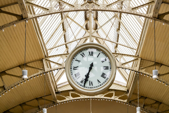 Large vintage clock hanging beneath an ornate glass and iron station roof. Historic architecture symbolizing travel, punctuality