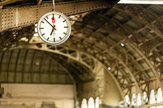 Round station clock hanging beneath an ornate iron railway hall roof. Travel schedule, punctuality, transportation