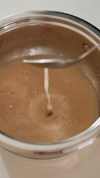 Overhead close-up of creamy brown sauce inside stainless steel pot as metal spoon stirs while thin liquid stream pours into center, forming splash, concentric ripples, bubbles, and glossy texture