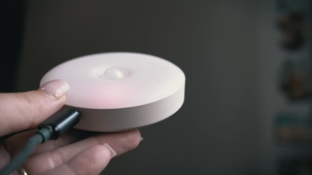 A hand holds a round rechargeable motion sensor LED night light plugged into a USB-C charging cable. Concept of smart home technology, energy-efficient lighting, and modern home automation.