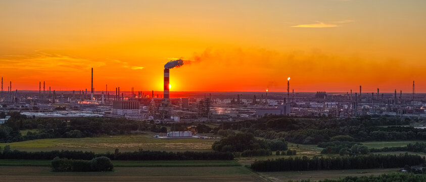 Aerial view of the PKN Orlen refinery with a smokestack against a vibrant sunset sky, surrounded by industrial infrastructure and fields in Plock, Masovian Voivodeship, Poland.