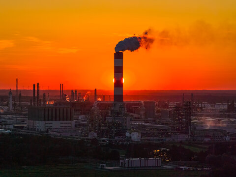 Aerial view of the PKN Orlen refinery at sunset with a tall chimney emitting smoke against a vibrant orange sky in Plock, Masovian Voivodeship, Poland.