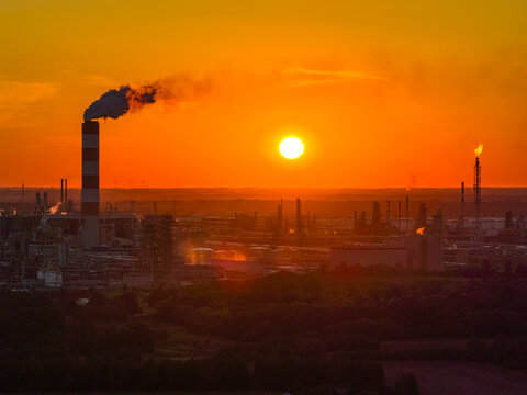 Aerial view of the PKN Orlen refinery with a prominent smokestack under a vibrant sunset sky, surrounded by industrial infrastructure and lush greenery in Plock, Masovian Voivodeship, Poland.