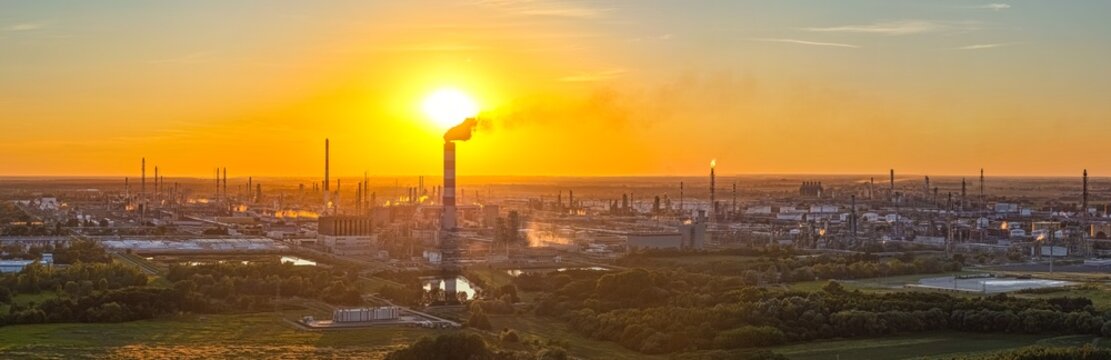Aerial view of the PKN Orlen refinery in Plock at sunset, with a tall smokestack, industrial structures, and green fields under a golden sky. Plock, Masovian Voivodeship, Poland.