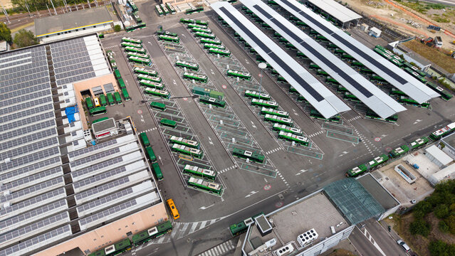 Aerial view of an electric bus depot featuring rows of parked green buses under solar panel roofs and urban infrastructure in Milan, Lombardy, Italy.