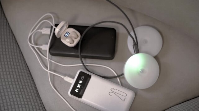 Portable power banks charge wireless earbuds and circular puck lights on a grey fabric surface. Concept of portable energy, modern electronic technology, and everyday battery recharging.