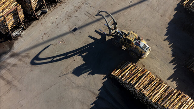 Aerial view of a log loader operating in a timber yard with stacks of logs and long shadows cast on the ground in Nelson, Nelson Region, New Zealand.
