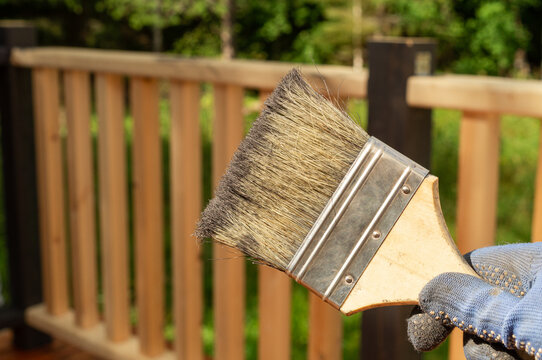 Hand in protective glove painting a wooden deck railing with a brush outdoors. DIY home improvement, deck maintenance and outdoor renovation concept.
