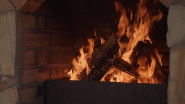 Close-up frontal view of active wood fire burning inside stone and brick fireplace, with tall flickering flames, glowing coals, stacked charred logs, rising sparks, warm amber light and rustic comfort