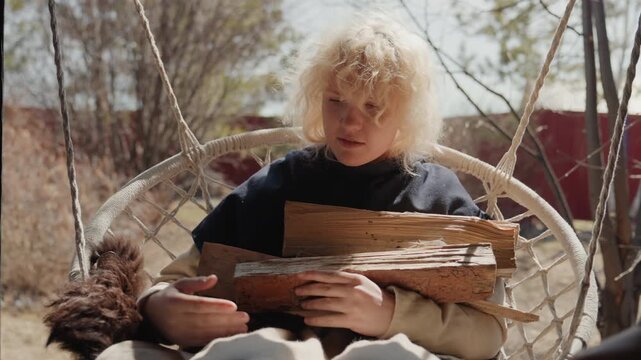 Curly blond child sits in woven hanging swing, arranging split firewood on lap and passing pieces to adult hands, with calm focused expression, rustic textures, soft daylight, outdoor family scene