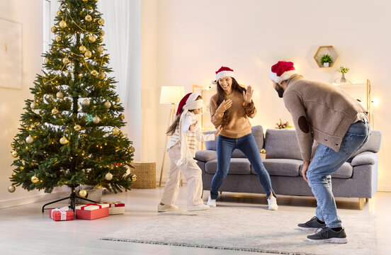 Christmas family playing hide-and-seek blindfolded with daughter at home. Parents and child girl in Santa hats play and having fun near festive tree by glowing lights. Winter holiday joy.