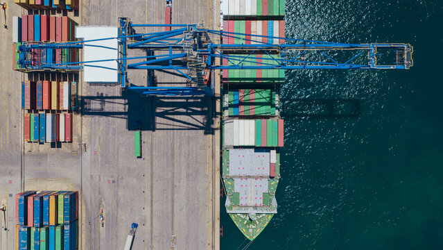 Aerial view of a large container ship docked at Piraeus Harbour, featuring colorful shipping containers, a massive gantry crane, and deep blue water in Athens, Greece.