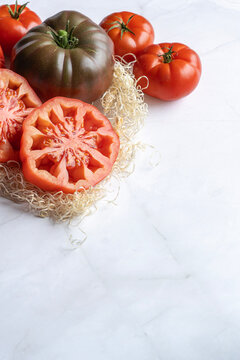 Fresh sliced red heirloom tomato with whole Kumato tomatoes on wood wool. Organic farm harvest for healthy cooking, Mediterranean diet, and recipes. High angle view with copy space on white marble bac