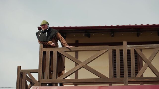Helmeted worker in high visibility vest leans over timber balcony railing while signaling toward lower level, hands gripping balustrade, body tilted forward during outdoor site communication