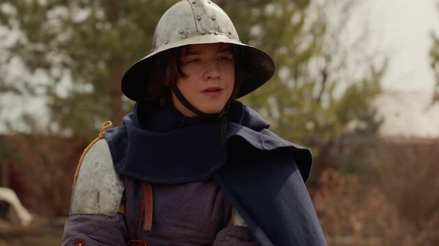 Medieval reenactor wearing layered armor and wide-brimmed helmet completes practiced sword movement, lowering blade with calm focused posture in outdoor courtyard beneath overcast sky, soft diffuse