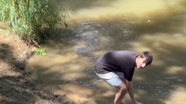 Happy teenage boy playing with his black dog in a muddy stream on a sunny day. Carefree moments of friendship and fun in nature